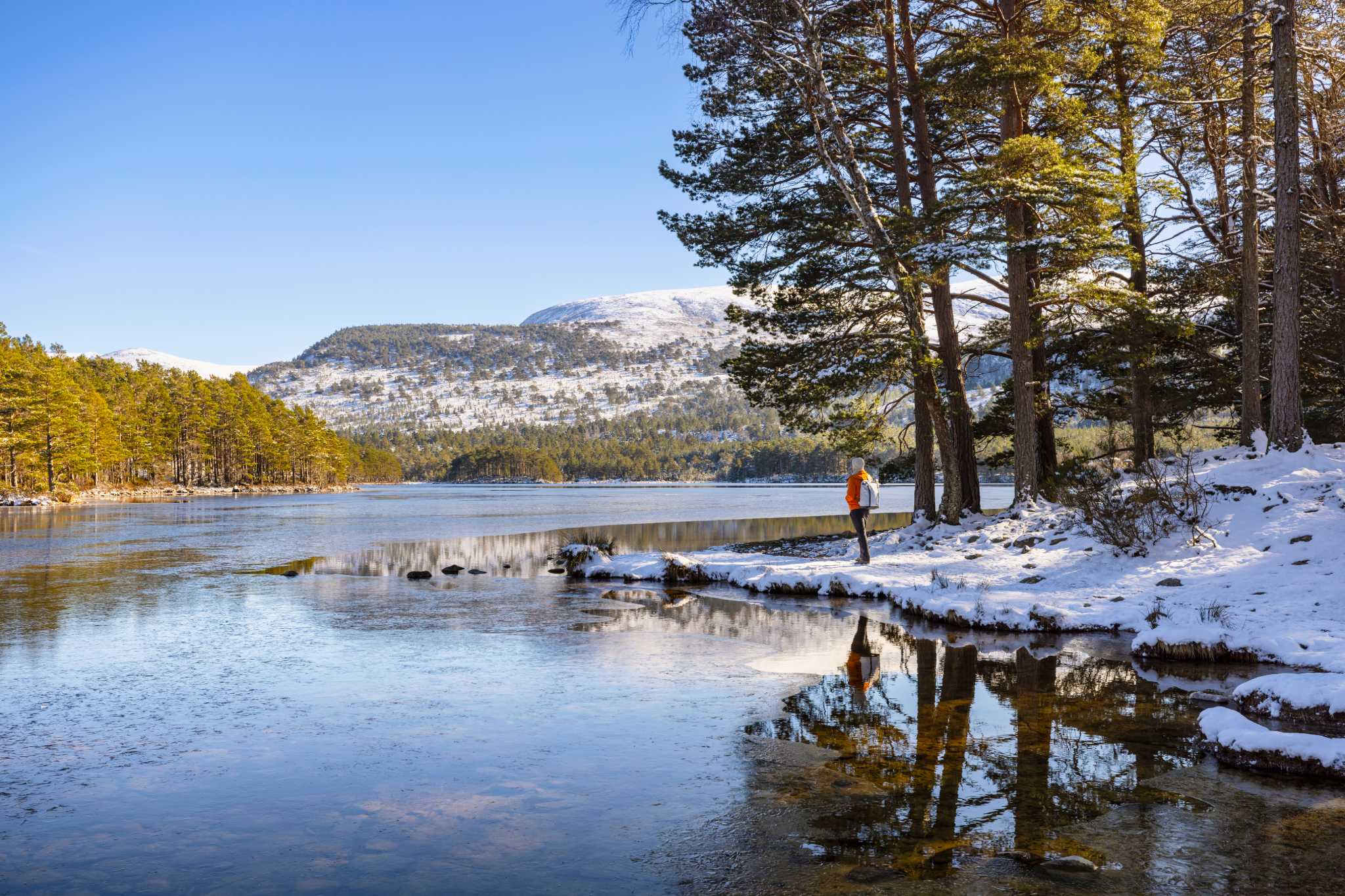 hiker stands by snowy banks of a loch with snow covered hills in the background