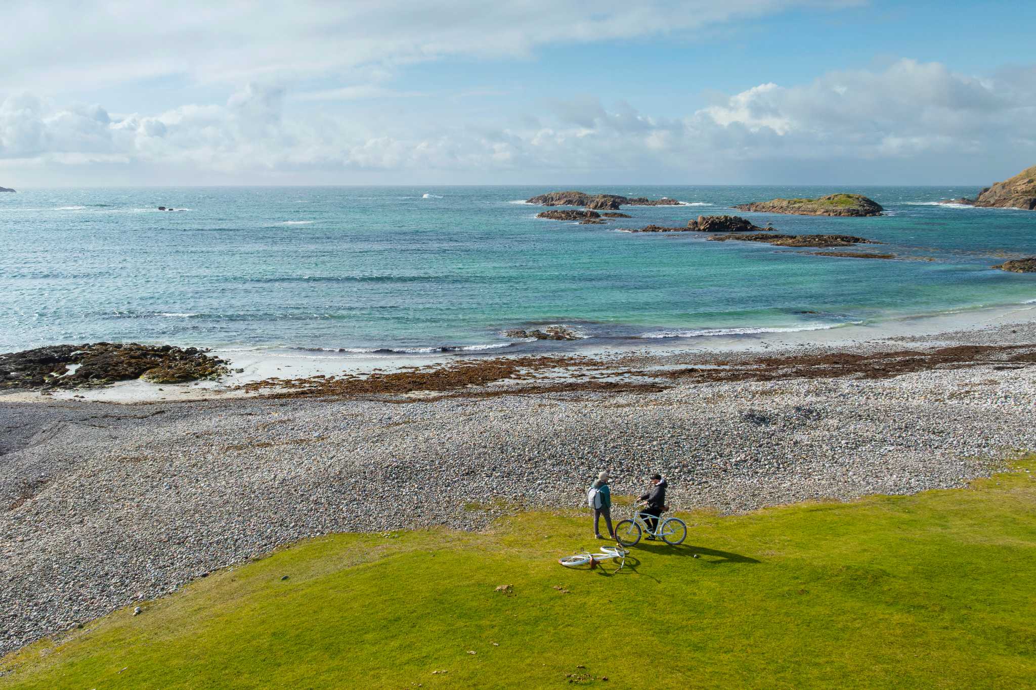 Two cyclists stand on a pebbled bay looking out towards sea on a sunny day.