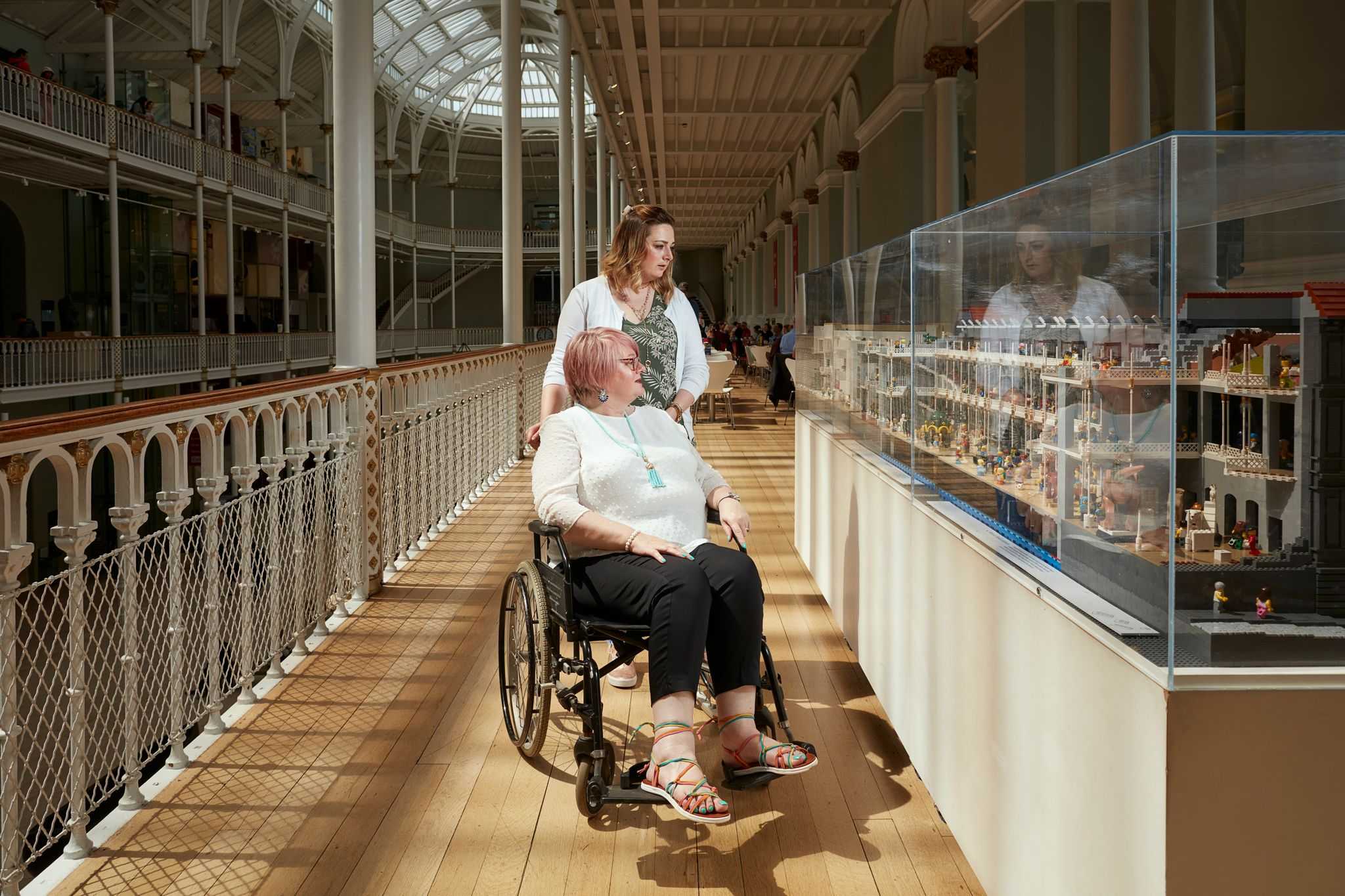 A woman standing and a woman in a wheelchair look at a museum exhibit.
