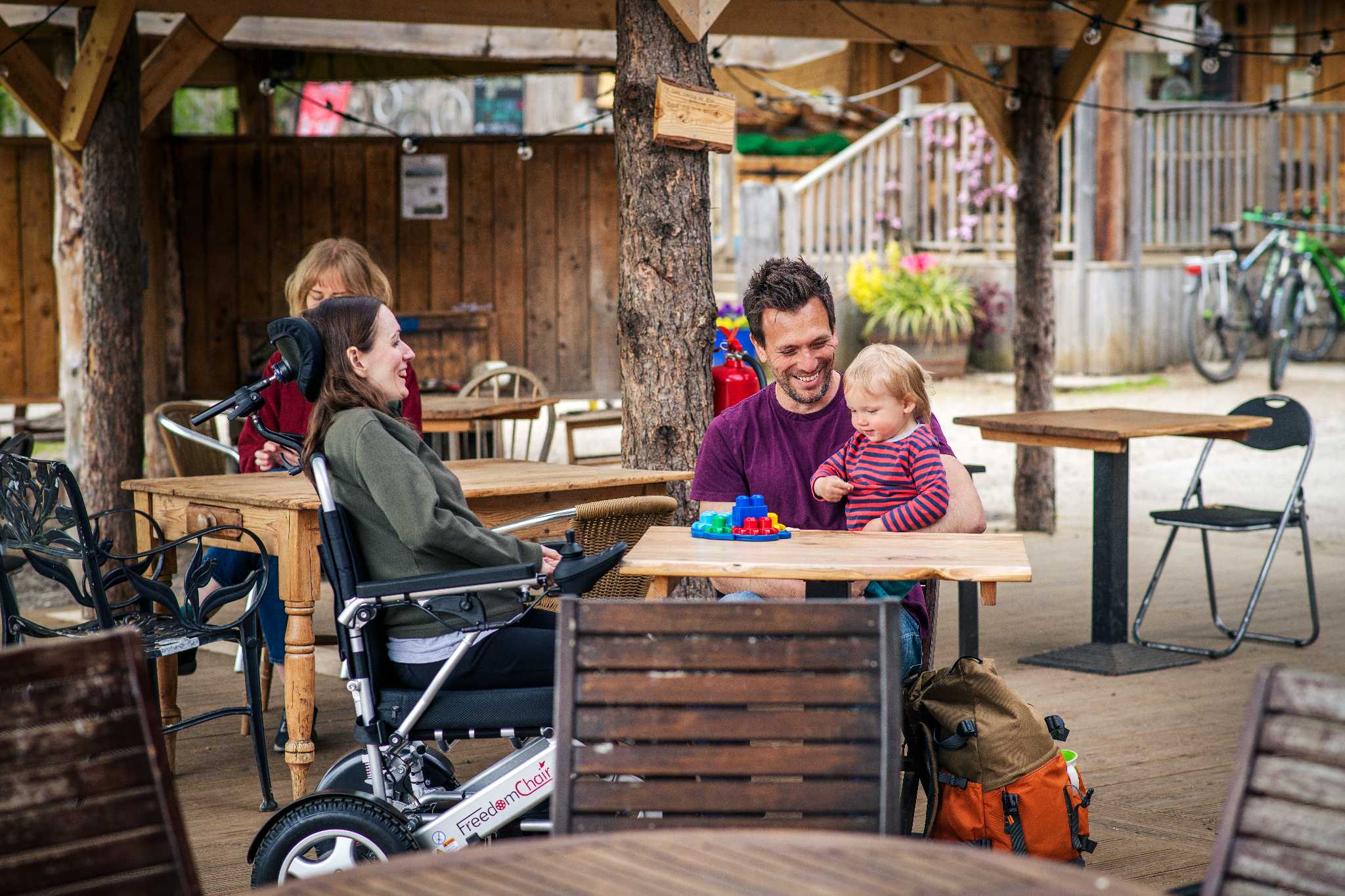 A woman seated in a wheelchair and a man with a small child sit at at exterior dining table smiling.