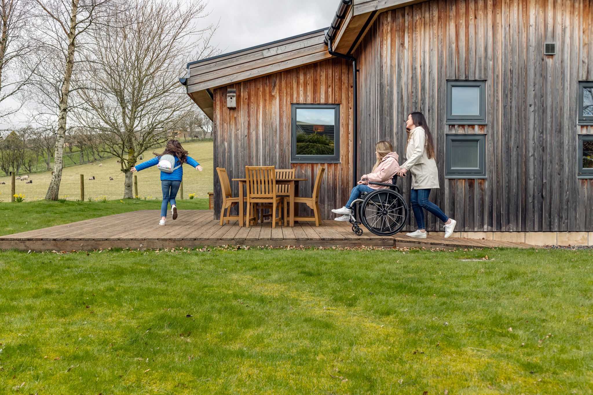 Exterior of wooden lodge, a girl in a wheelchair is pushed along decking surface while a younger child runs ahead