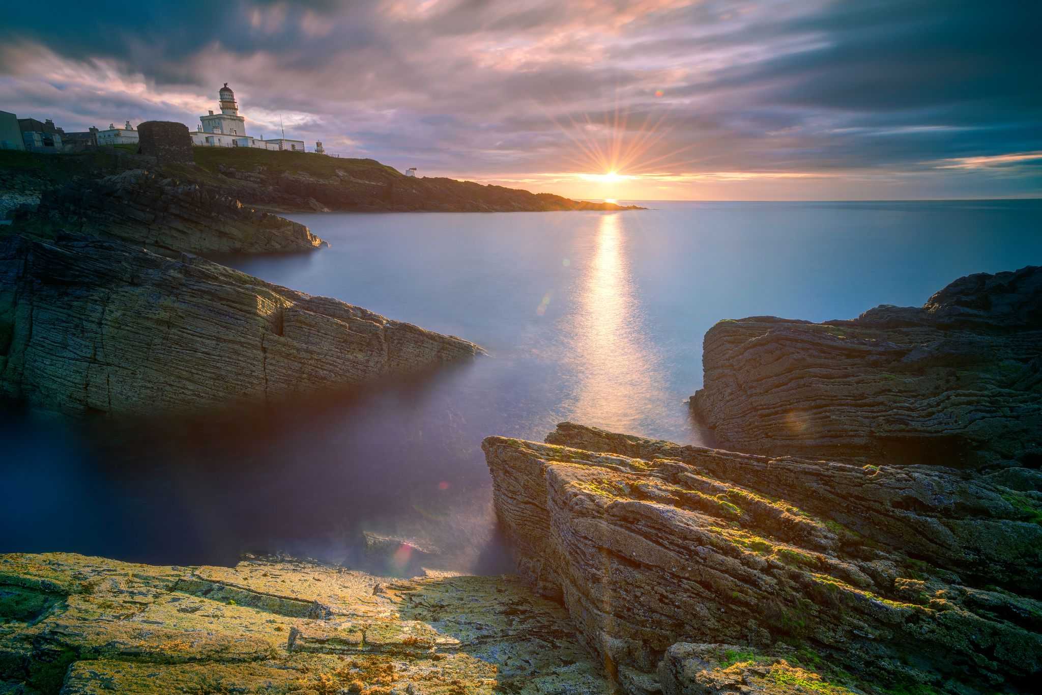Looking across a calm sea inlet to a lighthouse as the sun sets on the ocean's horizon.