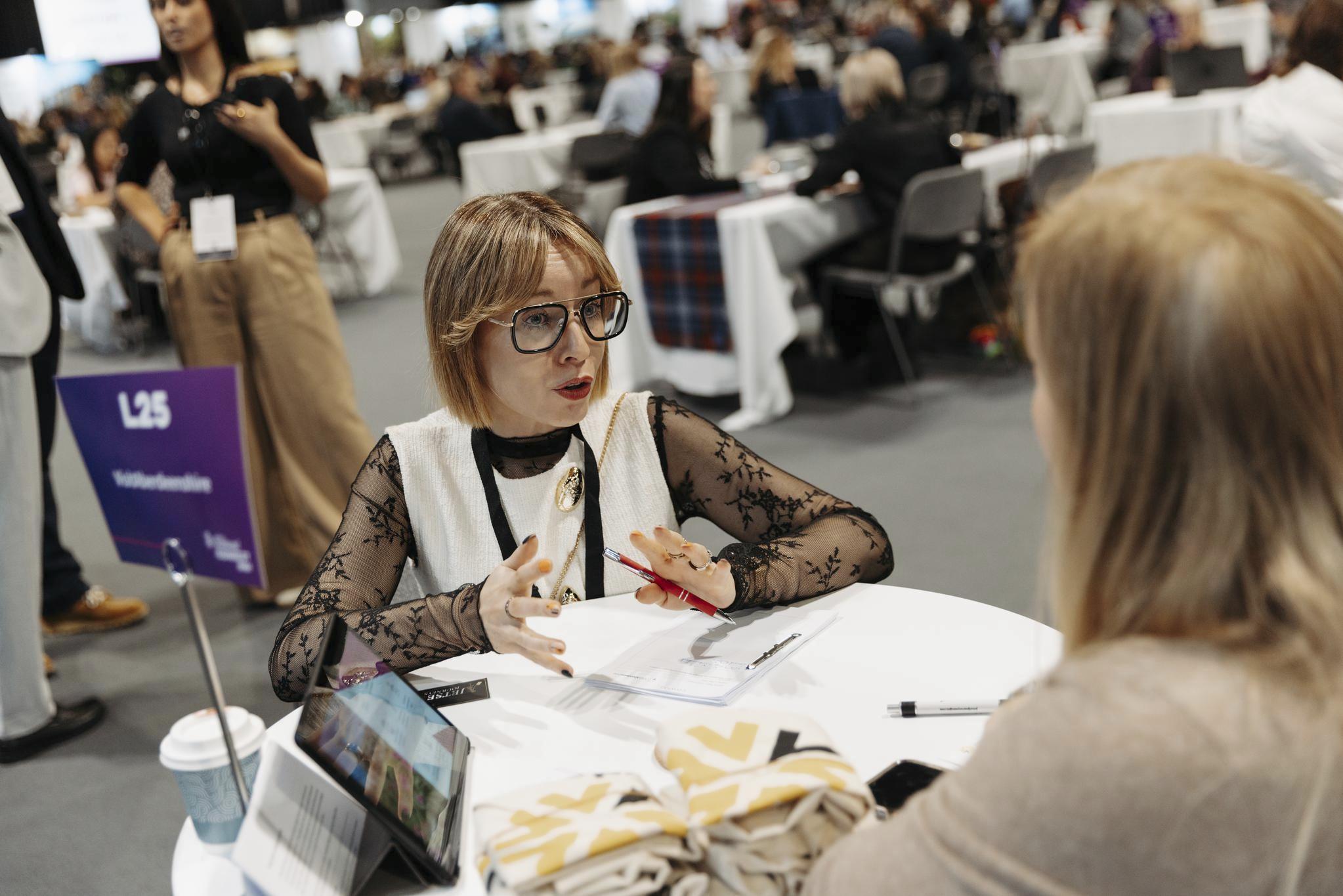 Two business professionals engage in conversation at a table at a busy conference venue.