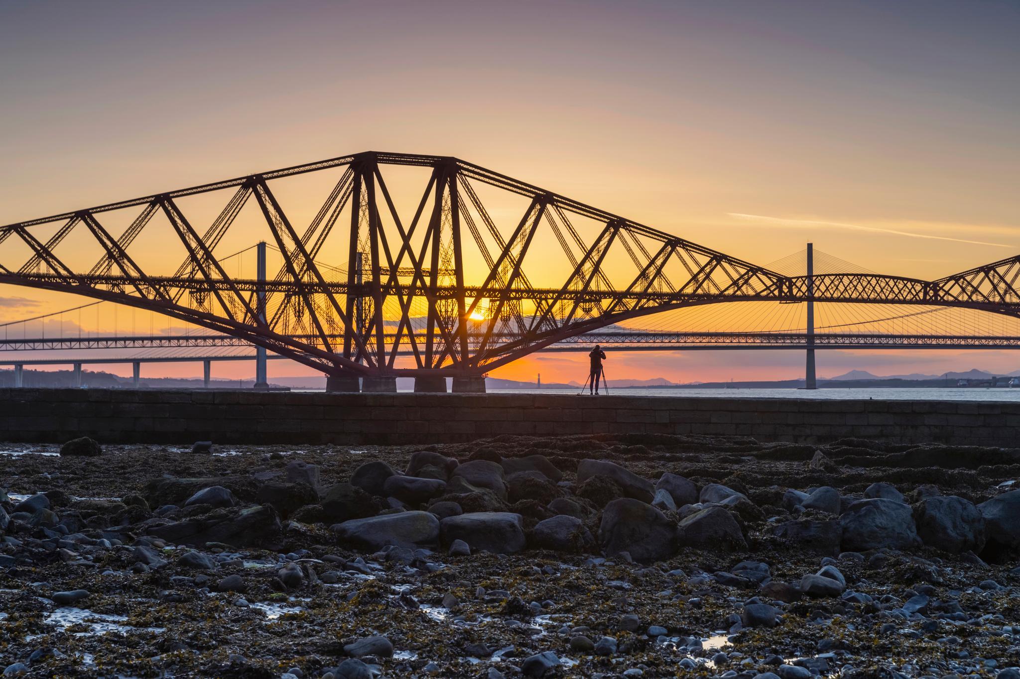 A photographer is silhouetted as the sun is setting behind the Forth Bridge.