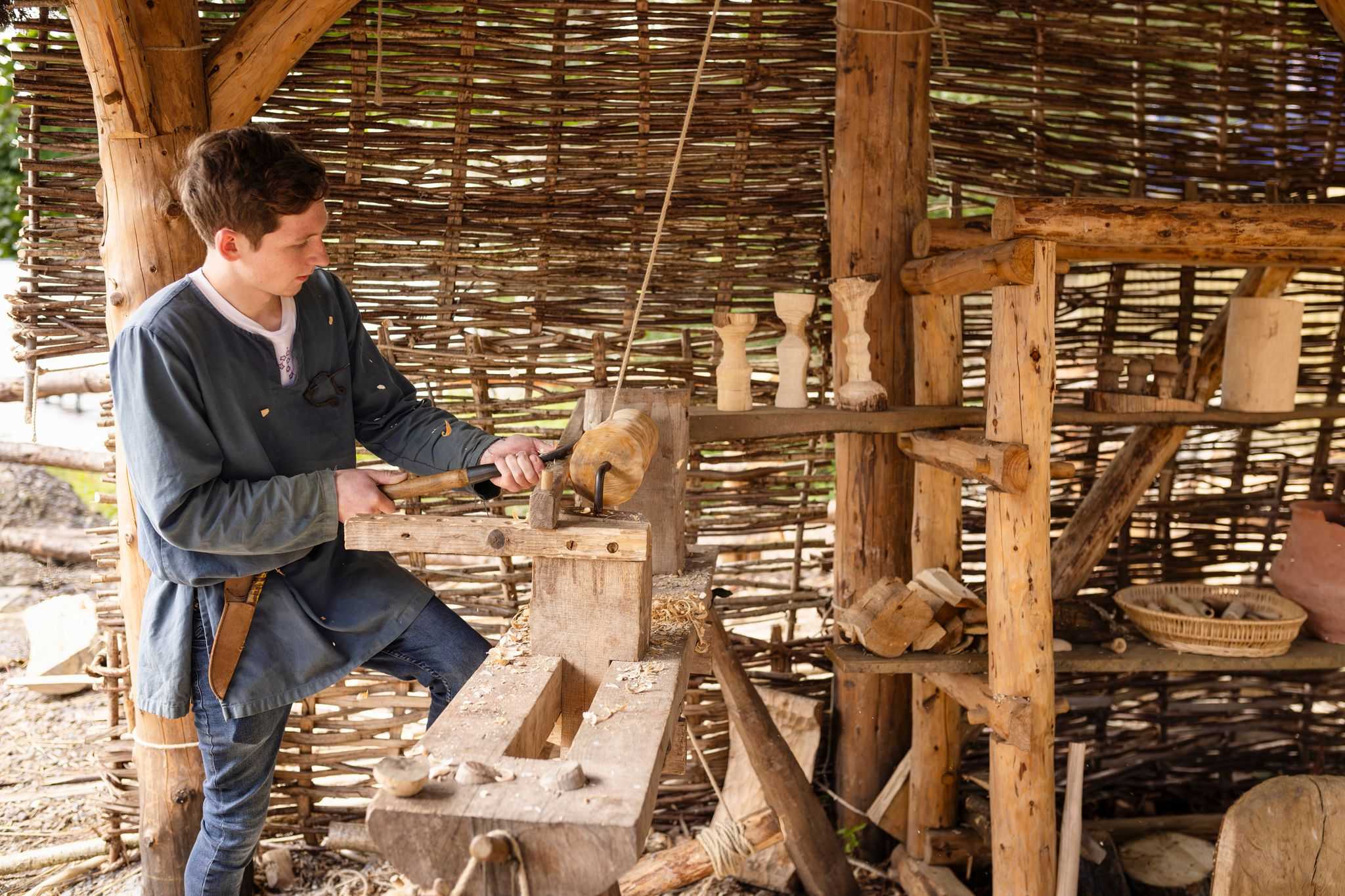 A young man in iron age costume carves wood on an iron age router