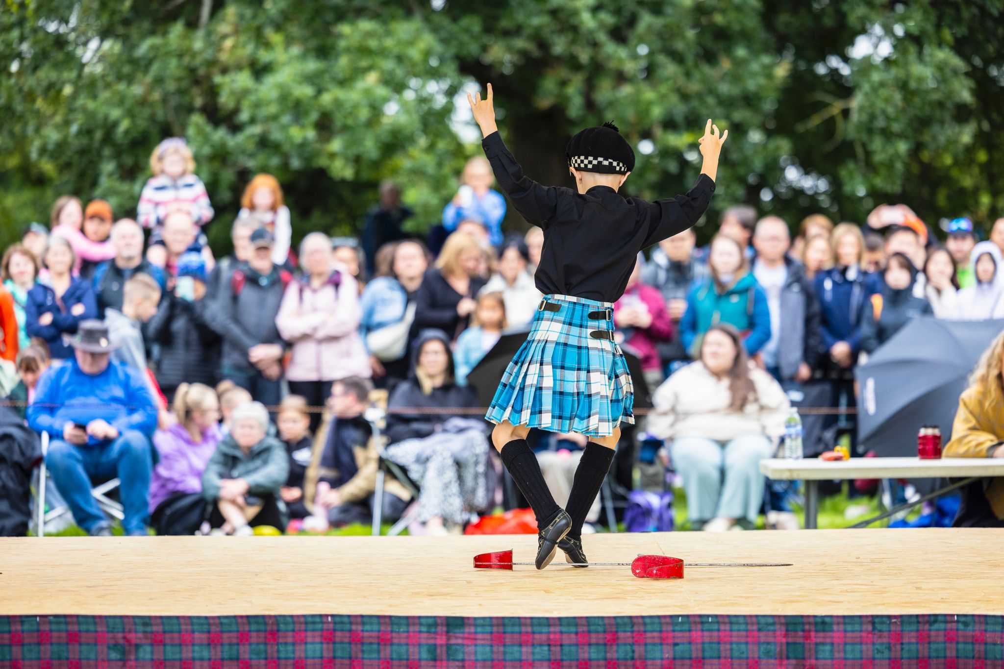 A young boy in a kilt does a dance over crossed swords on a stage with crowd watching