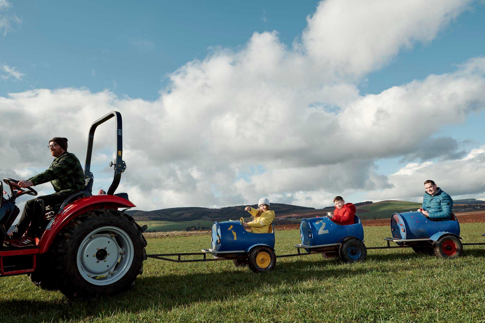 Two kids and an adult are enjoying being pulled along in trailers by a tractor