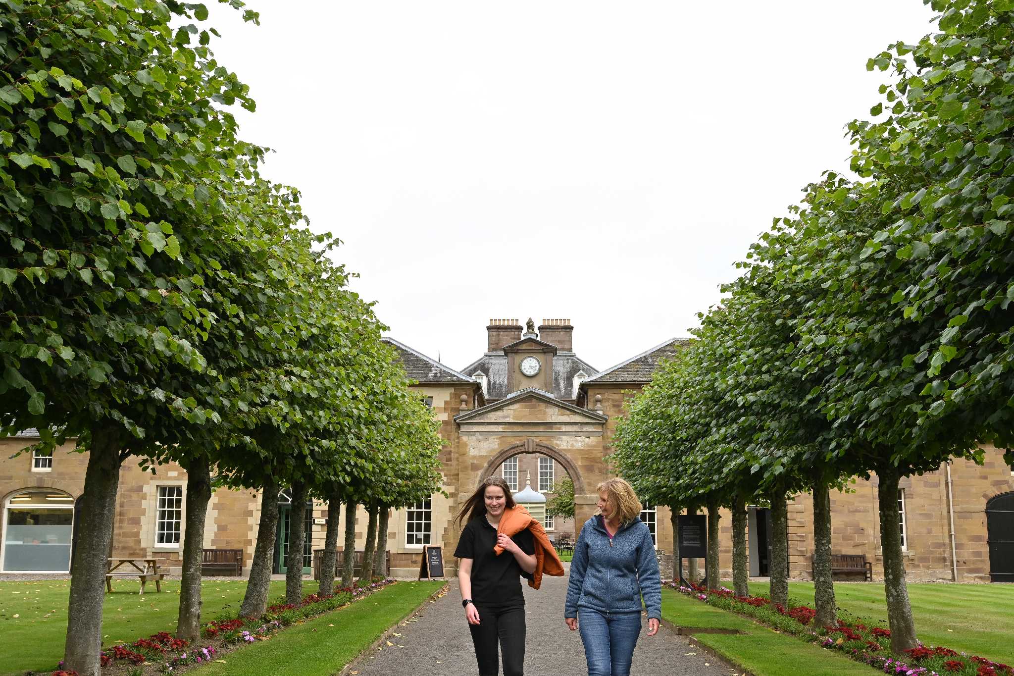 Two women walk along a tree lined pathway away from an arch way with a tower and clock face above it.