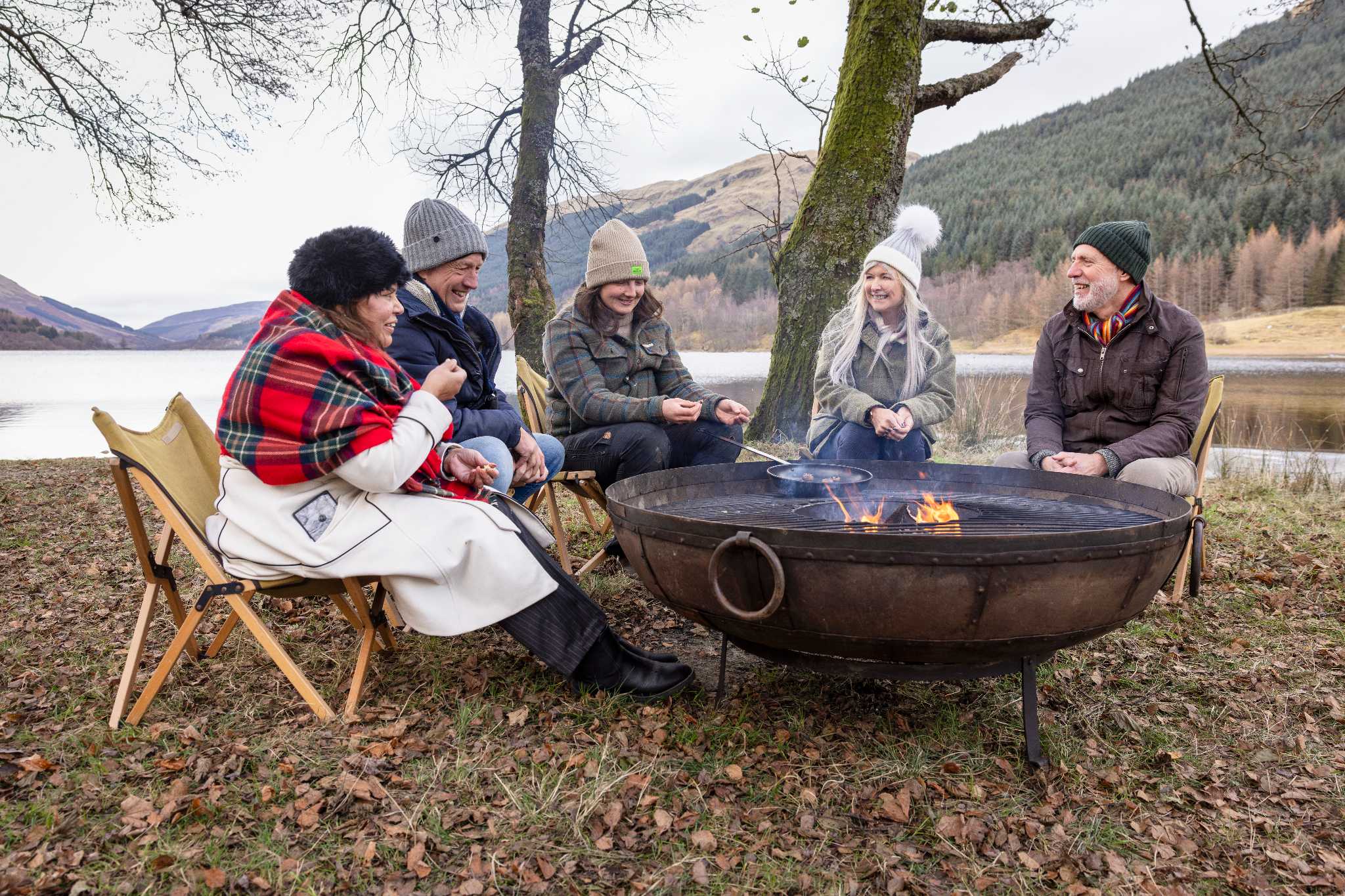 On a cold day, five happy people in warm clothes sit around a large barbeque, by a loch, as one of them cooks in a frying pan.
