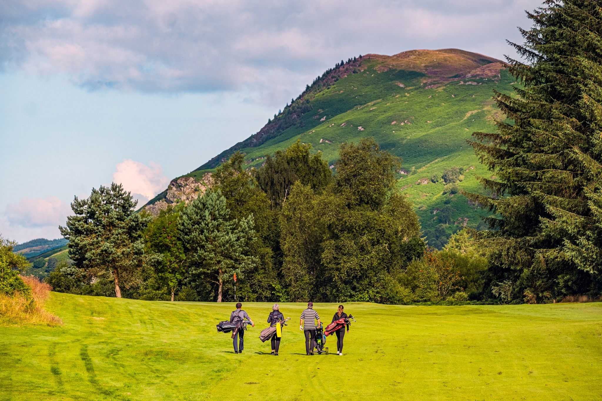 Four figures with golf bags walk across a green course. There is a large hill in the background.