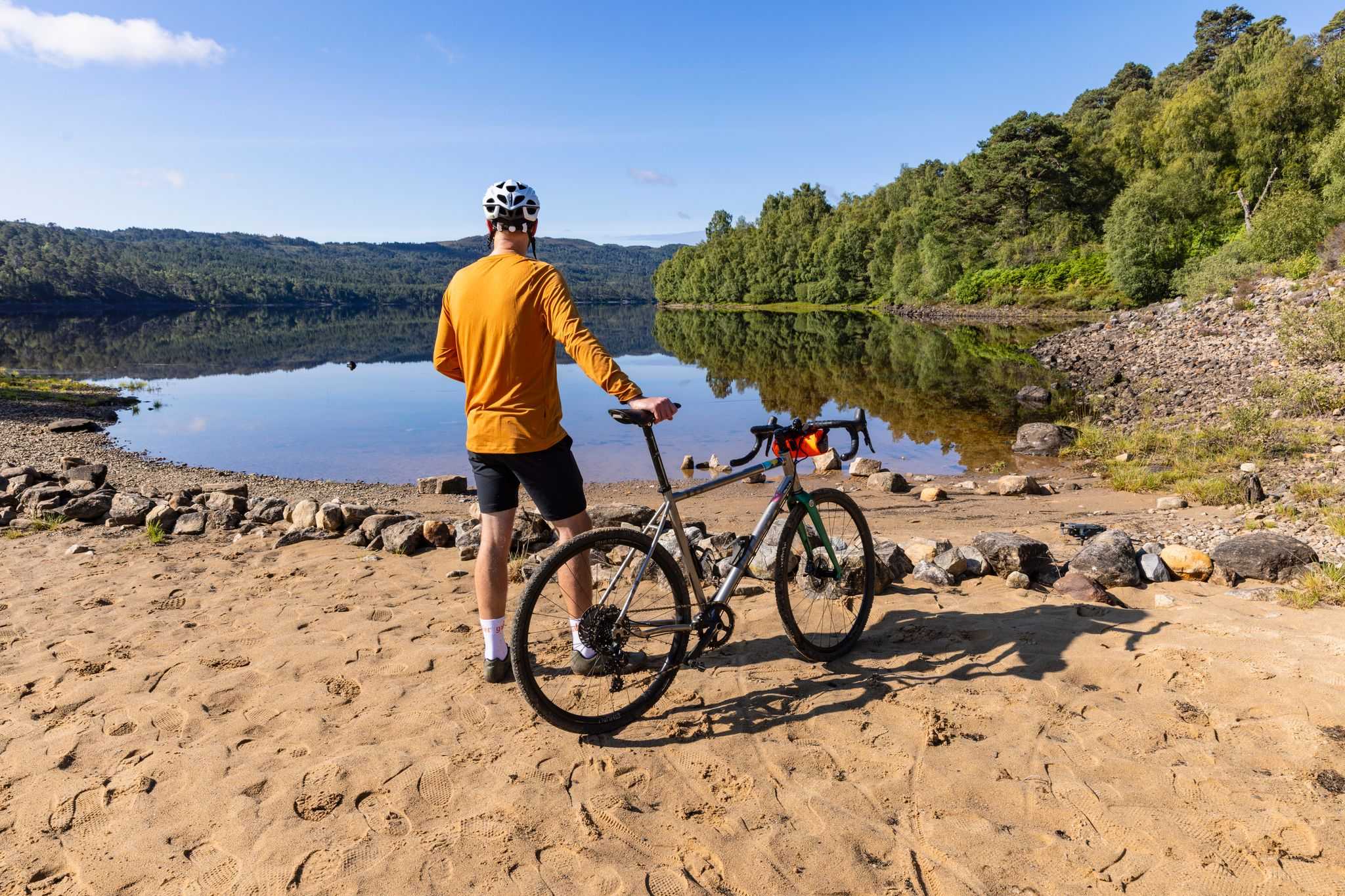 A cyclists stands with his bike at the shore of a loch on a sunny day