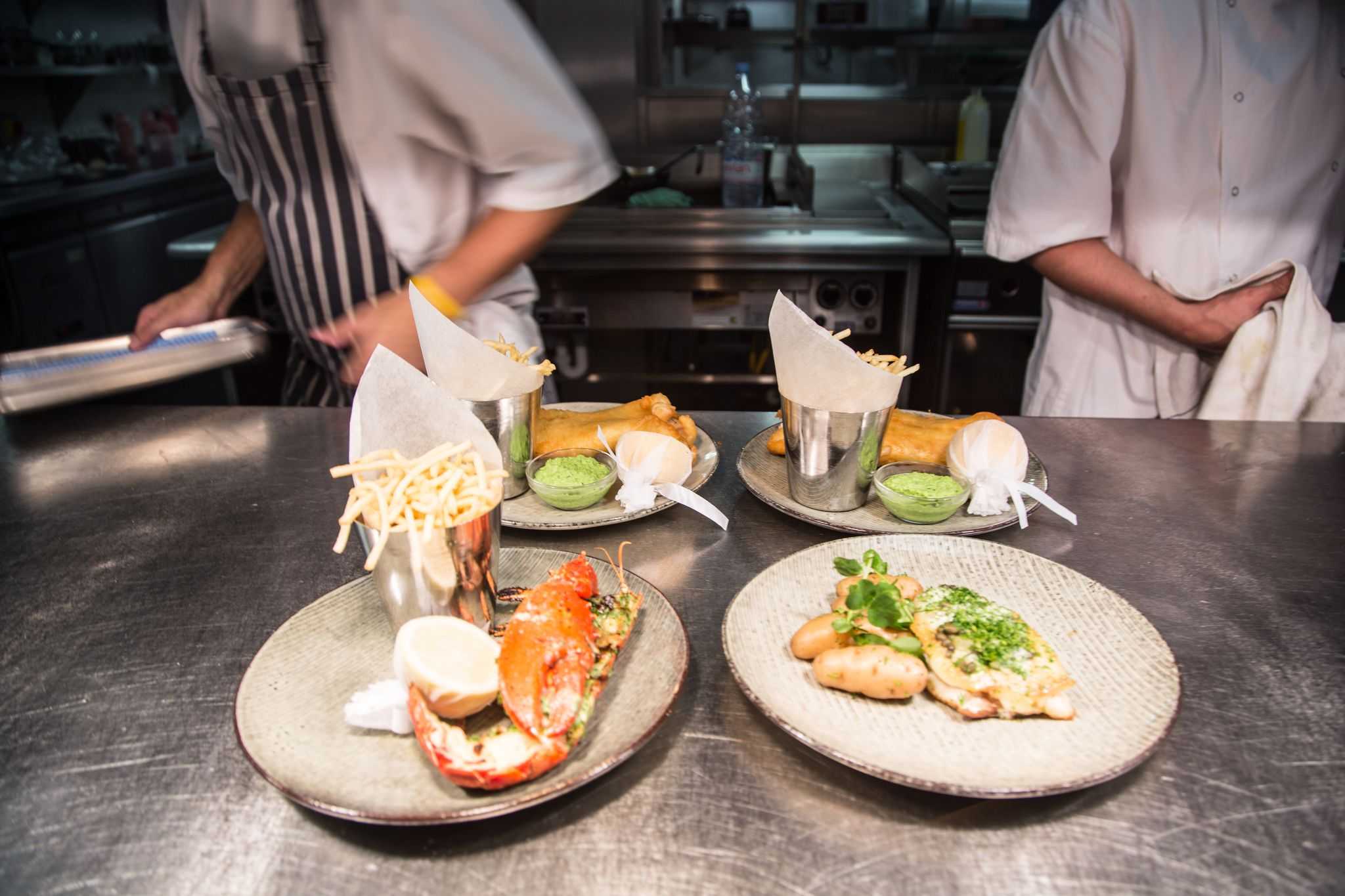 The image shows a restaurant kitchen with plated dishes on a counter. Two chefs in white uniforms are visible in the background.