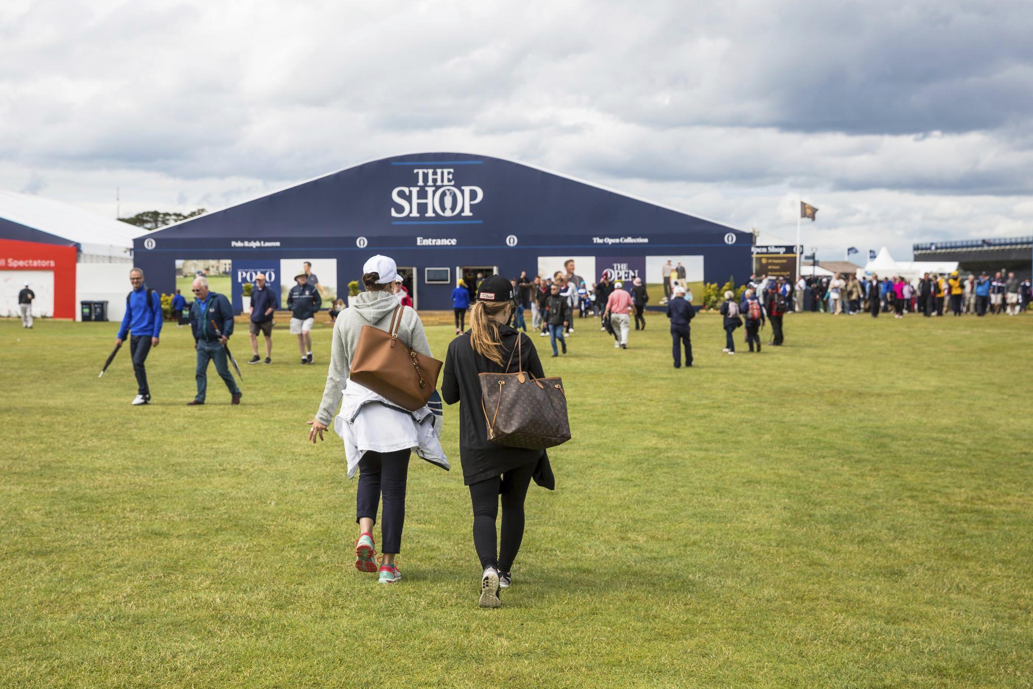 Two women walk across a grass field towards a large shop at the Open golf event.