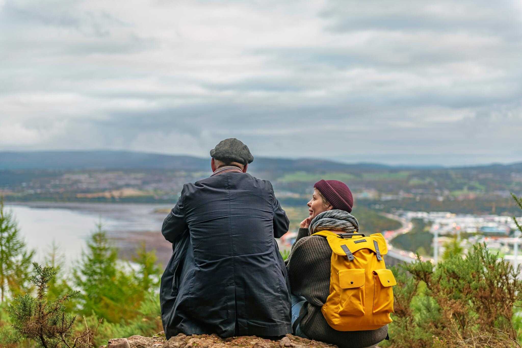 A couple sit on a hill overlooking trees and a large body of water with hills in the distance