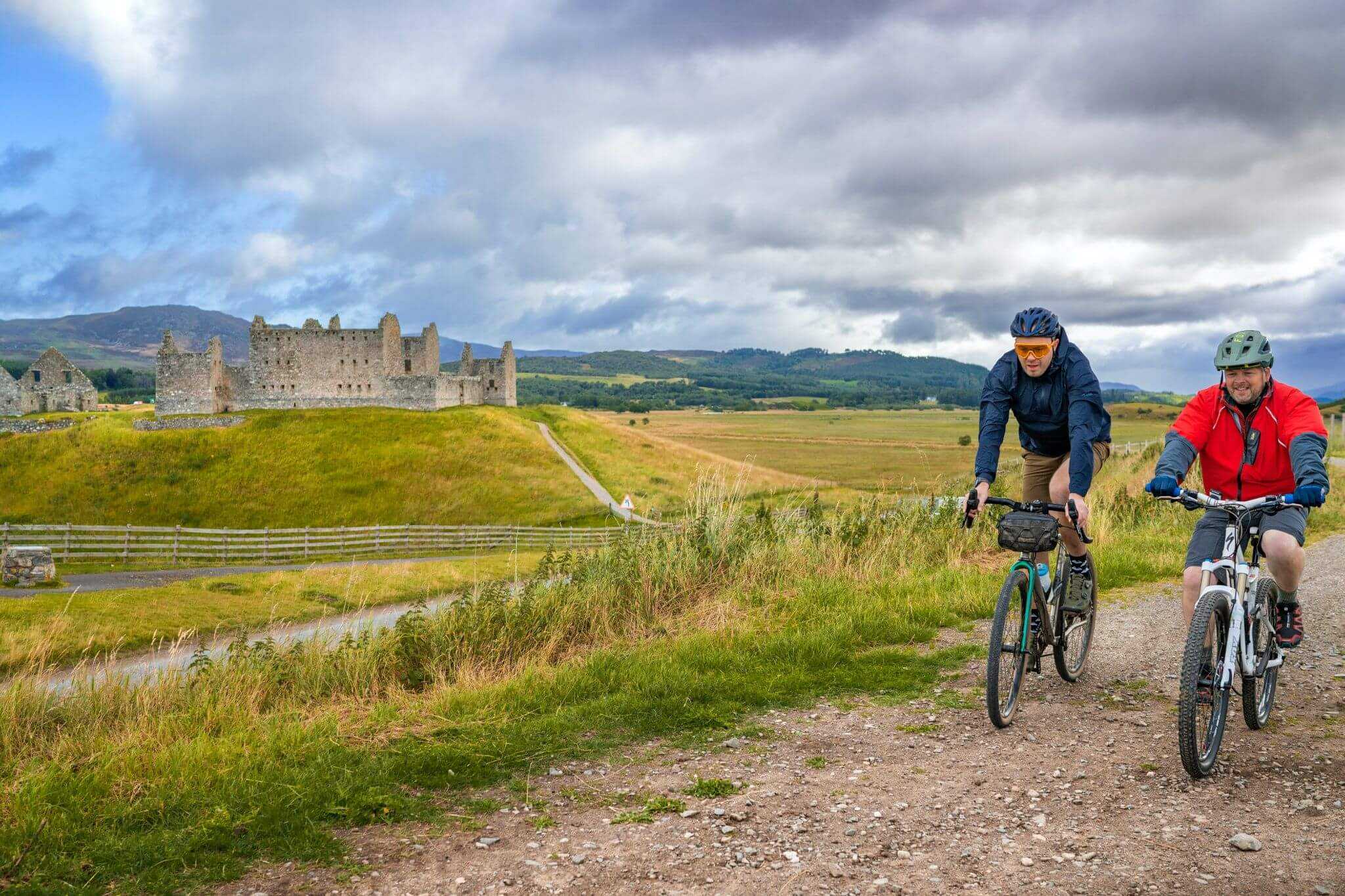 Two cyclists on a track cycle past large ruins of a building