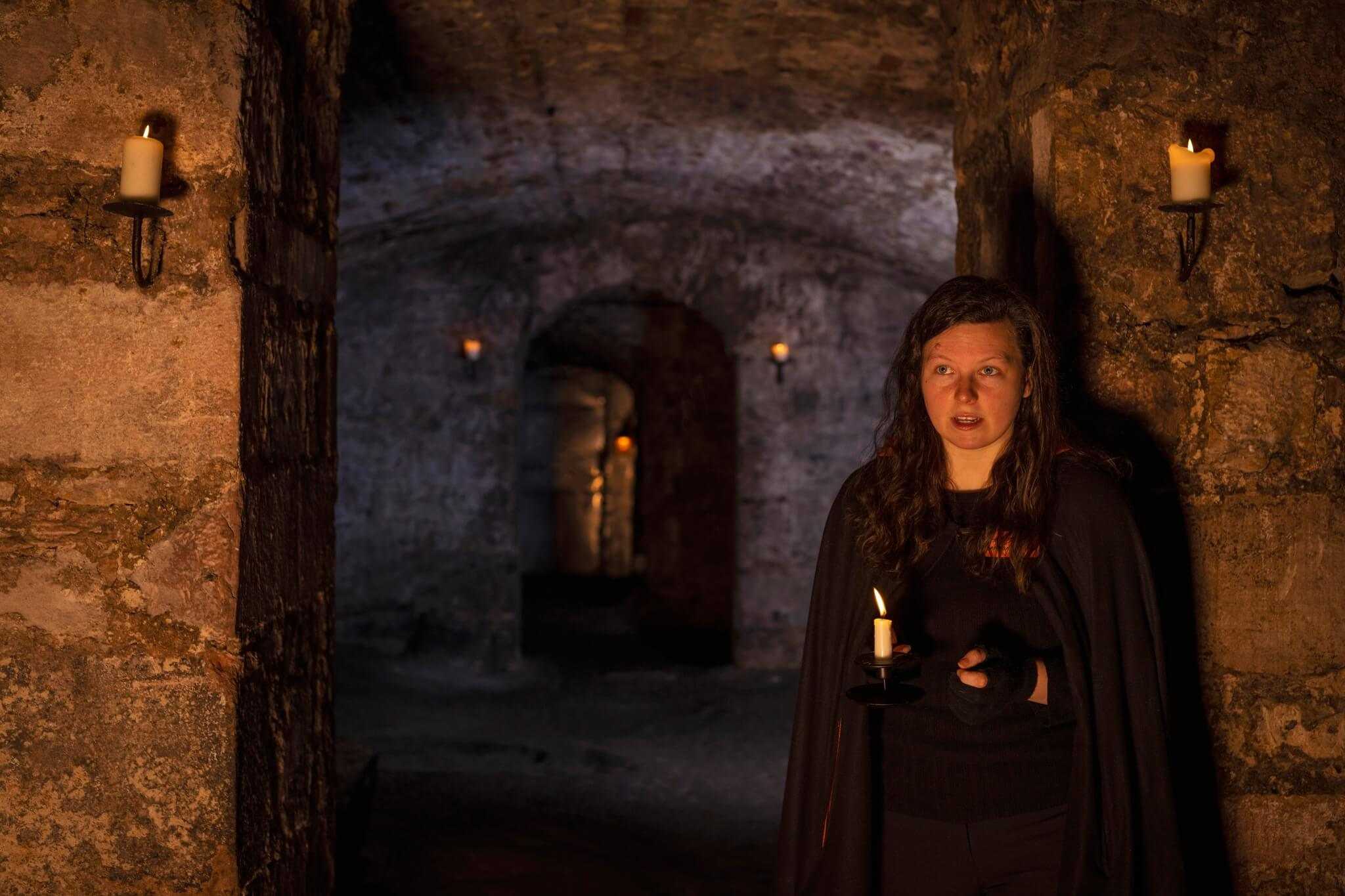 A tour guide in a long cloak stands in a dark vault lit by two candles