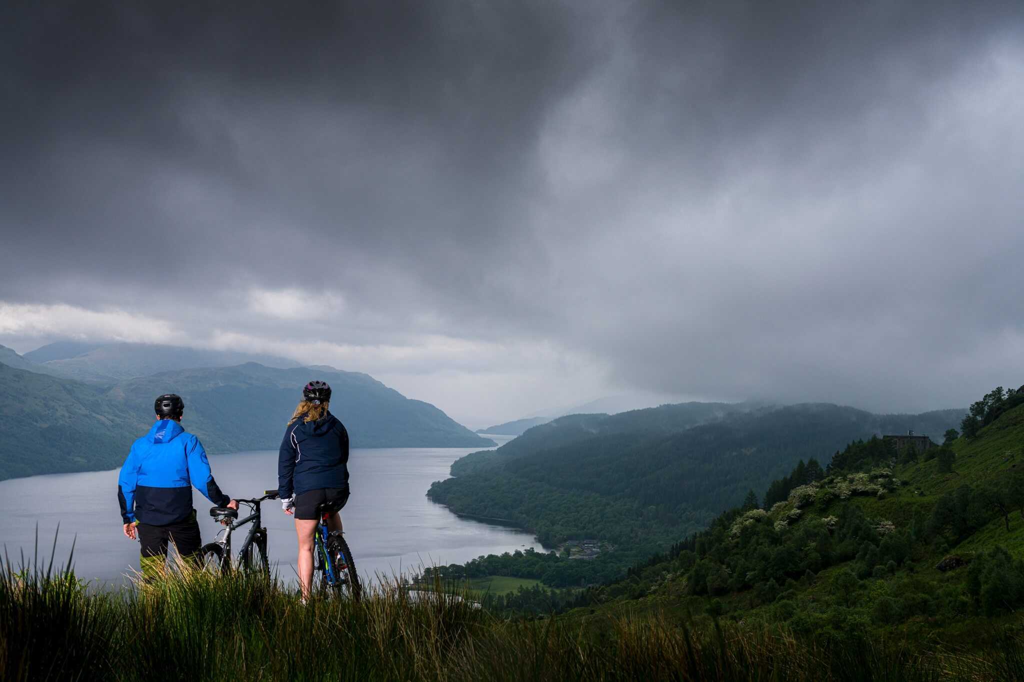 A couple with mountain bikes look down from a hill onto Loch Lomond