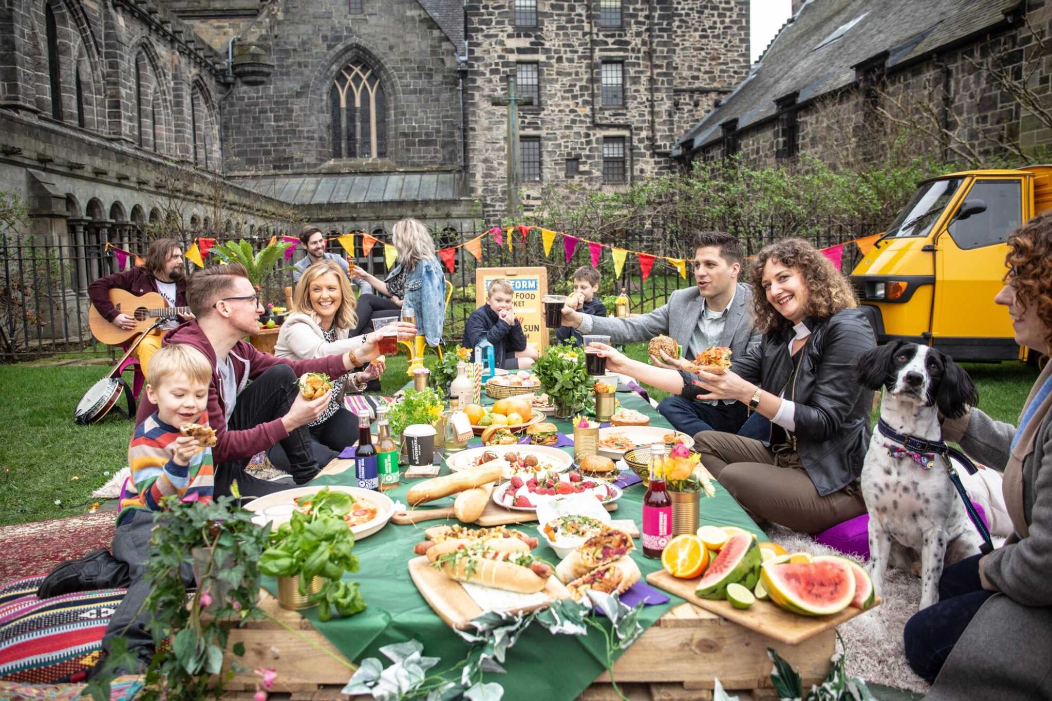 families sit outside around a long table laden with food while a guitarist sits in background