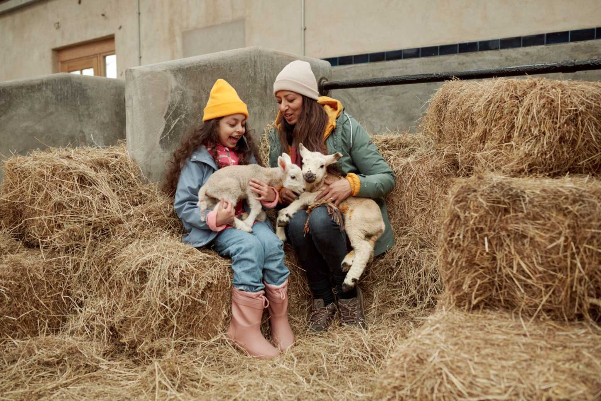 A mother and daughter sit on straw bales bottle feeding young lambs