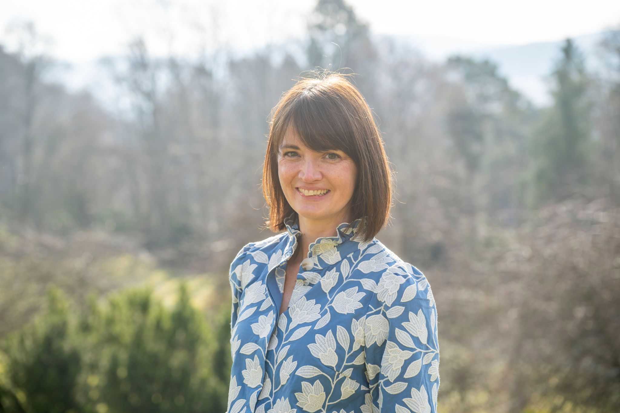 A lady stands in a wooded area smiling at the camera