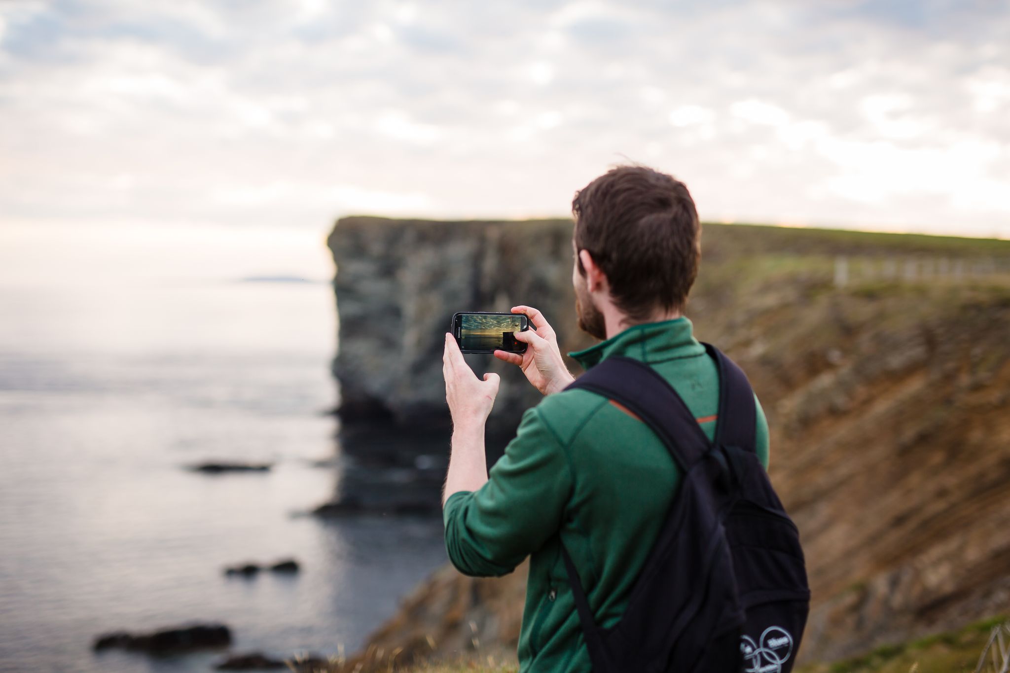 A man stands at the edge of a cliff taking a photo with his mobile