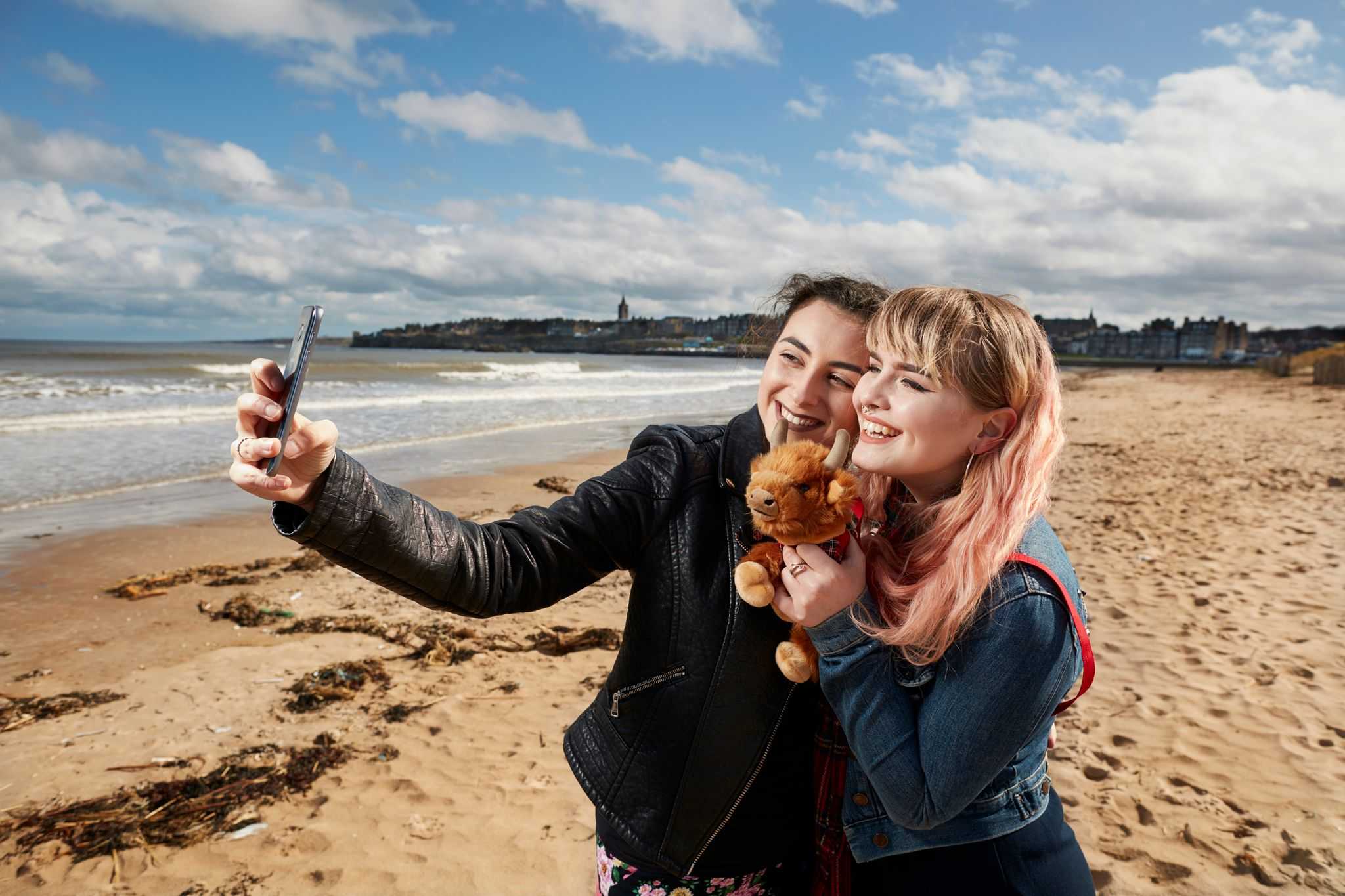 Two young women take a selfie on a beach. the tide is out and buildings are visible in distance