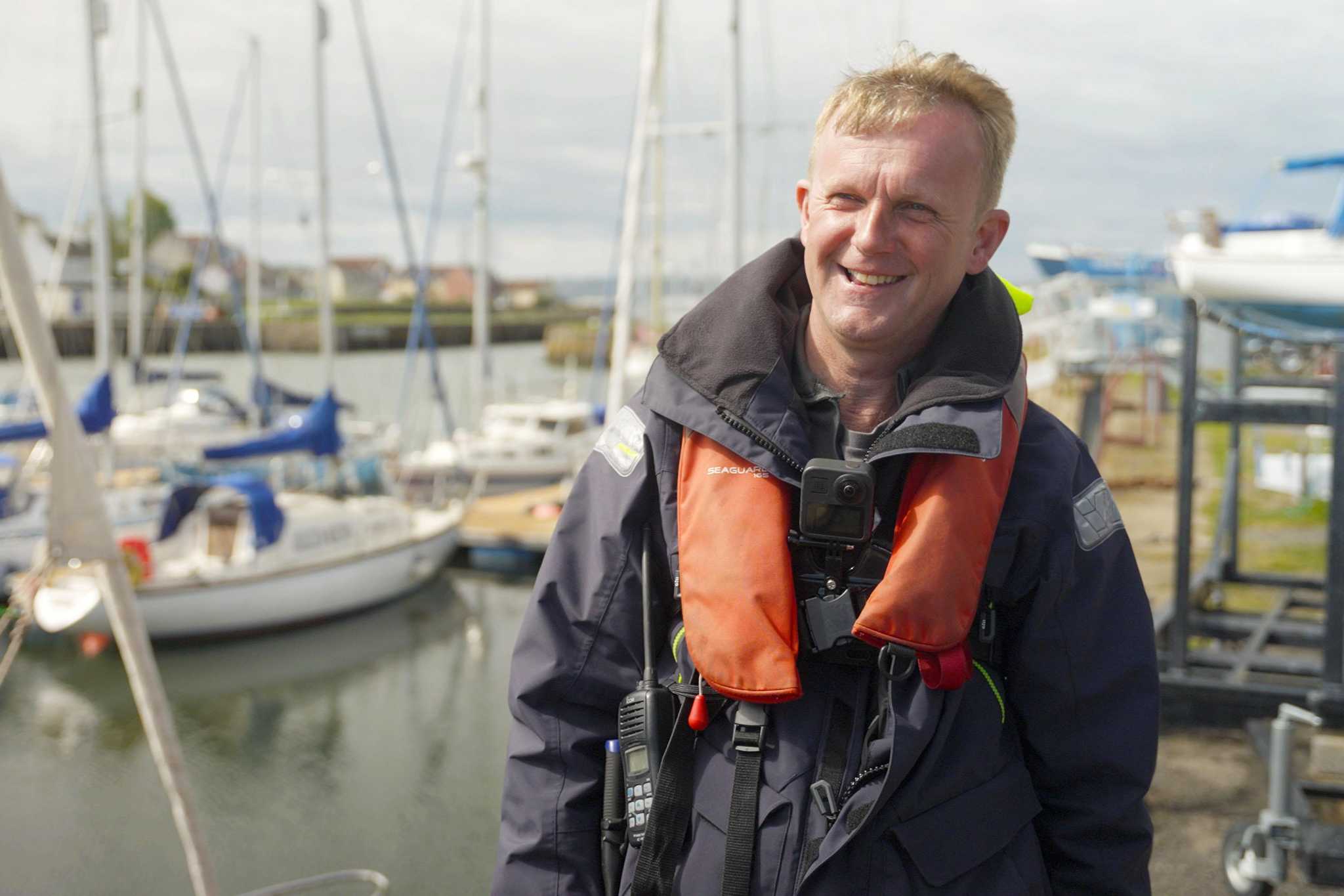 A man in a life vest stands smiling in a marina. Boats can be seen moored in the background.