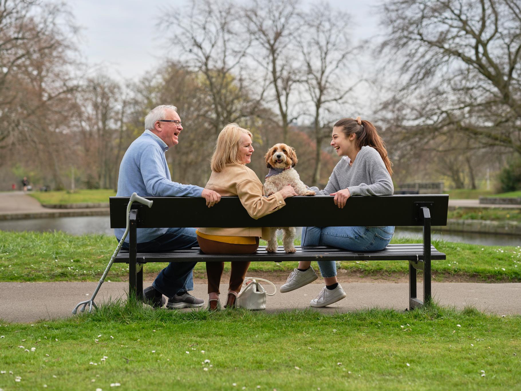 An elderly couple relax with a dog and a younger lady on a bench in a park